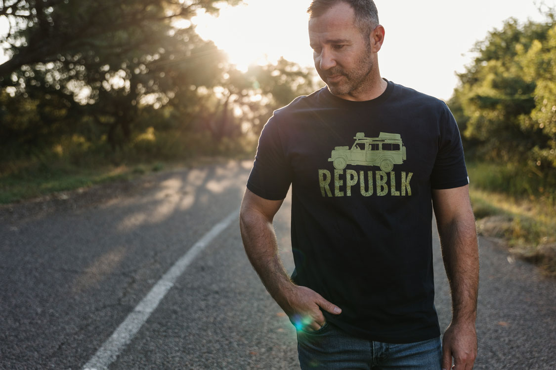 Man in a navy REPUBLK graphic tee on a country road at sunset